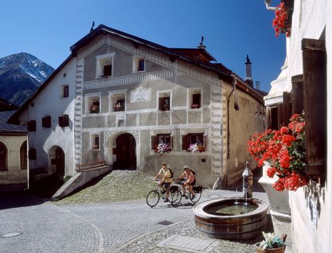 Deux cyclistes passent devant une maison traditionnelle et une fontaine à Bever. 