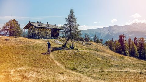 A mountain biker rides towards an old stone house at the top of a hill.