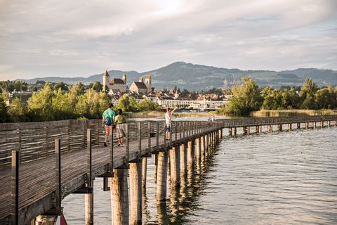Einige Menschen laufen auf der Holzbrücke, die über dem See liegt. Im Hintergrund sieht man eine Burg.