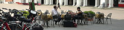 Auf dem Palazzo hat es ein paar Tische und Stühle, 2 Touristen sitzen dort und trinken etwas. Links vom Bild stehen viele Fahrräder.