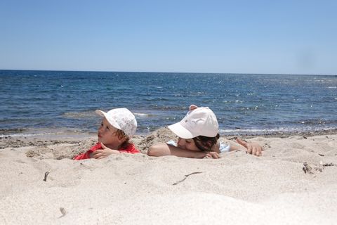 Zwei Kinder mit Käppi sind im Sand eingebudelt.