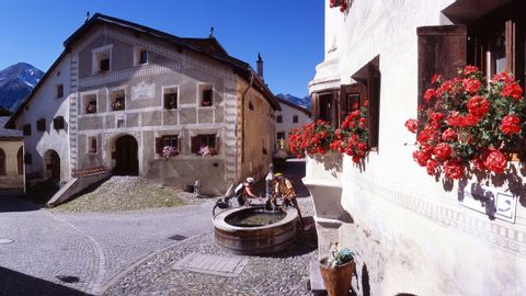 Two trail runners refresh themselves at the village fountain between the typical Engadine houses. The windows are decorated with geranium pots.