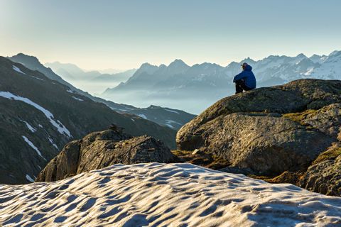 Person sitzt auf Felsen und schaut sich das Panoramadach der Berge an. Auf den Bergwiesen liegt noch Schnee