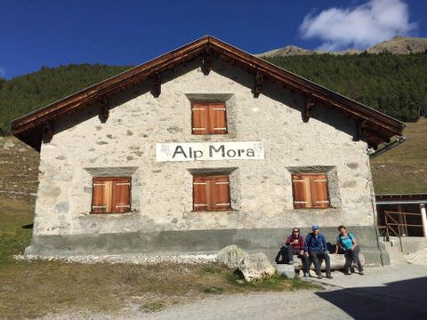 Eine Alphütte mit geschlossenen Holzländen und der Aufschrift "Alp Mora". Auf einer Bank vor dem Haus machen 3 Wanderer Pause