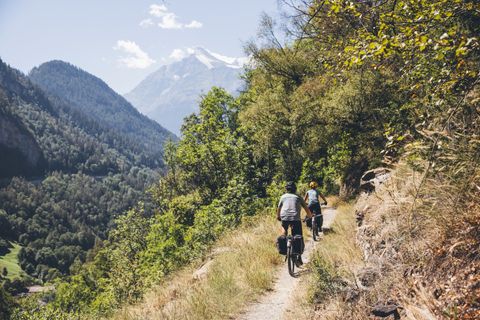 Des cyclistes roulent l'un derrière l'autre sur un étroit chemin de montagne en Valais. En arrière-plan, des sommets de montagne blancs. 