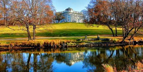 Ein Herrschaftliches weisses Gebäude im Hintergrund, dass sich im See vorne am Bildrand spiegelt, umgeben von einer Wiese mit Bäumen in den schönsten Farben des Herbstes.