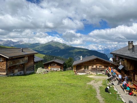 Auf einer grünen Wiese stehen drei Holzhäuser auf der Alp Medergen auf der Mittelbündner Panoramaweg Wanderung. 