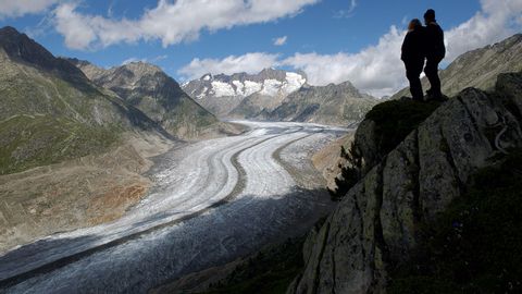 Zwei Wanderer stehen erhöht auf der Bergkante und blicken auf die Gletscherzunge des Aletschgletscher.