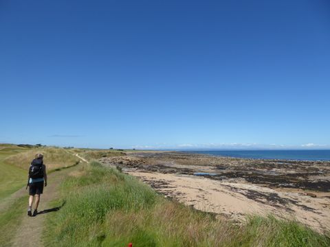 Links im Bild eine Wanderin die auf einem schmalen Naturpfad der Küste auf der linken Seite, spaziert. 2/3 des Bildes sind mit einem strhlend dunkelblauen Sommerhimmel gefüllt.