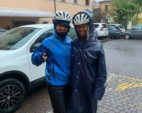 Zwei Frauen posieren vor einem Auto in Regenjacken und Fahrradhelmen. 