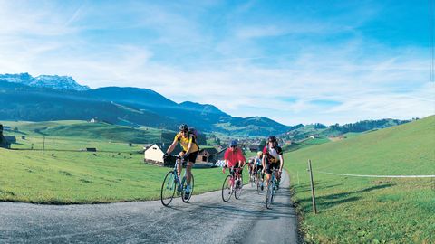 A group of racing cyclists riding up a side road, with a beautiful Alpine panorama in the background.