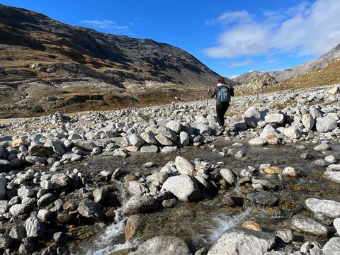 Eine Wanderin überquert einen Bach oder Fluss der mit Steinen belegt ist. 