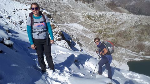 Zwei Wanderinnen stehen bei strahlender Sonne und kurzen Ärmeln im frischen Schnee auf dem Berg.