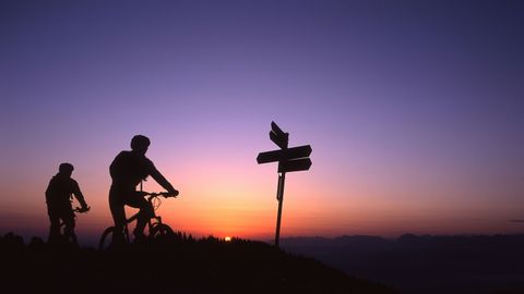 Deux vététistes passent à vélo devant un panneau indicateur au coucher du soleil.