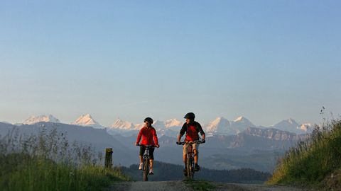A couple of bikers ride comfortably along a road in front of a panorama of snow-covered mountain peaks.