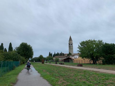 Auf einem Radweg neben einer Wiese fährt eine Velofahrerin in Regenjacke. Auf der rechten Seite ist ein Steinturm zu sehen. 