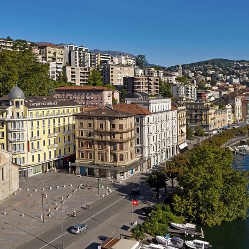 Luftaufnahme der Uferpromenade von Lugano mit Hotel International zwischen bunten Gebäuden am See, Bergen im Hintergrund, klarer blauer Himmel.
