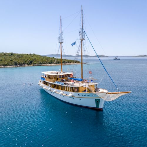 Weißes Motorsegelschiff MS Macek mit zwei Masten vor Anker in türkisfarbenem Adriatischem Meer nahe grüner kroatischer Küste.