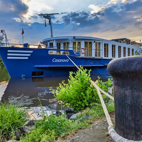 Blau-weißes Flusskreuzfahrtschiff MS Casanova am Anleger mit Poller im Vordergrund und grüner Vegetation am Flussufer.
