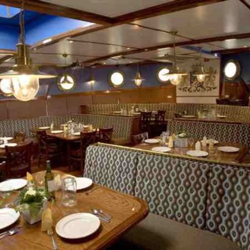 Innenraum des Mare fan Fryslan Restaurants mit Holztischen, gemusterten Banksitzen, Messingpendelleuchten und nautischem Dekor.