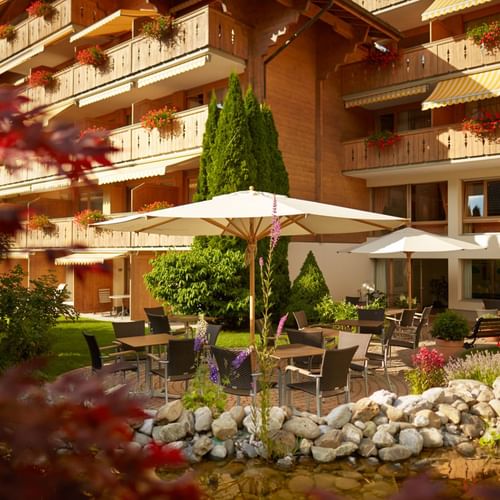 Wooden alpine hotel with flower-decorated balconies, garden terrace with white umbrellas, outdoor seating, and decorative pond in Gstaad.