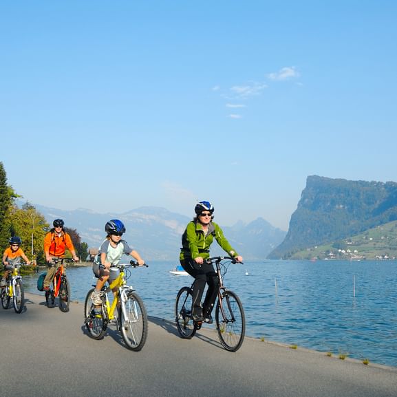 Vierköpfige Familie radelt auf Uferstraße am Vierwaldstättersee mit Bergen im Hintergrund. Klarer blauer Himmel und ruhiges Wasser sichtbar.