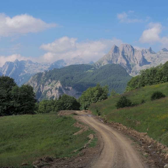 Unbefestigter Wanderweg bei Cerem durch grüne Hügel mit dramatischen Bergspitzen im Hintergrund unter blauem Himmel mit weißen Wolken.
