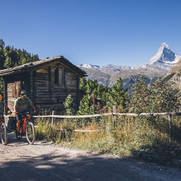 Deux cyclistes avec vélos sur une route de gravier près d'un chalet en bois, avec le Cervin emblématique visible en arrière-plan sous un ciel bleu clair.