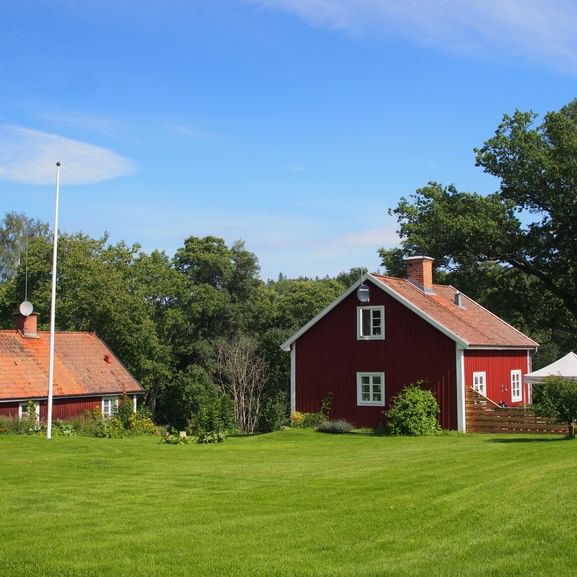 Zwei traditionelle rote Holzhäuser mit orangefarbenen Ziegeldächern auf grünem Rasen, umgeben von Bäumen unter blauem Himmel bei Mariefred.