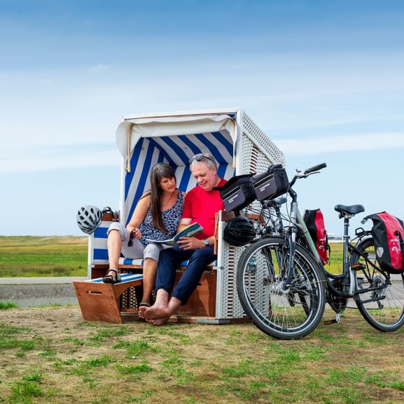 Zwei Radfahrer sitzen in einem blau-weiß gestreiften Strandkorb mit ihren Tourenrädern in der Nähe auf einer Wiese an der Nordsee.