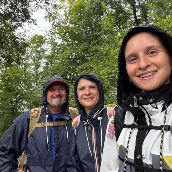 Drei lächelnde Wanderer in Regenjacken mit Kapuzen stehen in einem grünen Wald. Sie tragen Rucksäcke auf der Via Alpina in der Ostschweiz.