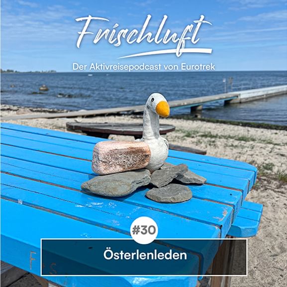 Blaue Holzbank mit Steinskulptur einer weißen Gans am Küstengewässer. Text zeigt 'Frischluft' Podcast Folge 30 über Österlenleden.