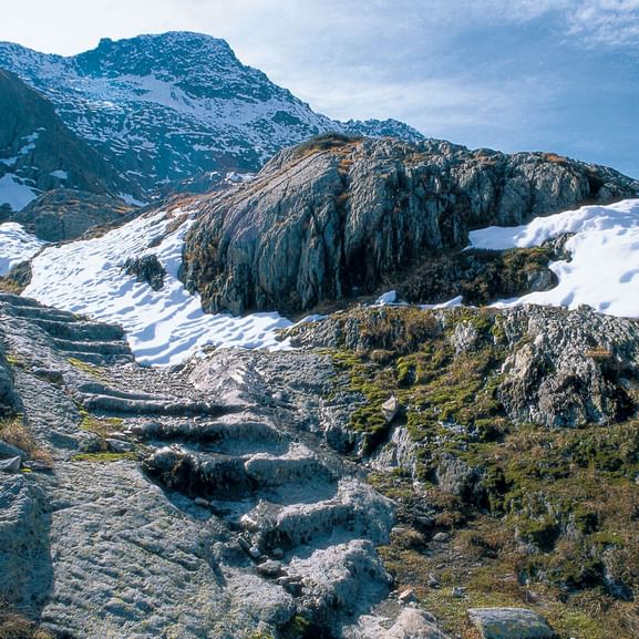 Von der Mitte nach links ist eine in den Fels gehauene Treppe, die an Steinhügeln und mit Schnee bedeckten stellen, vorbei zum Berg führt. Via Francigena. Wanderferien mit Eurotrek.