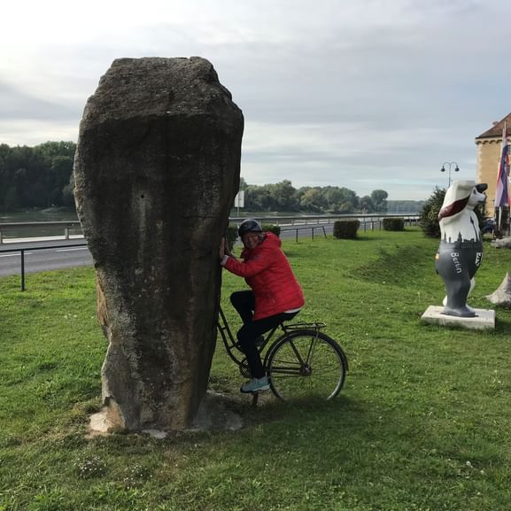 Prisca in roter Jacke mit Fahrrad lehnt sich an hohen Steinmonument, mit dekorativem Berliner Bär auf Gras in Mauthausen.