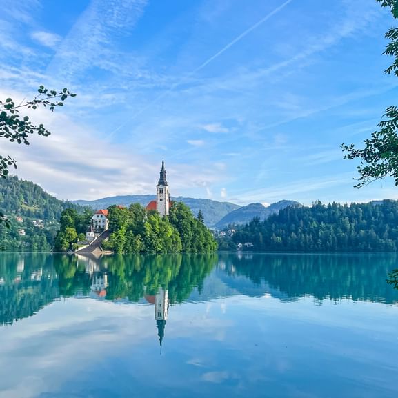Église avec clocher sur petite île boisée du lac de Bled, Slovénie. Reflet parfait dans l'eau calme, entourée de montagnes et arbres.