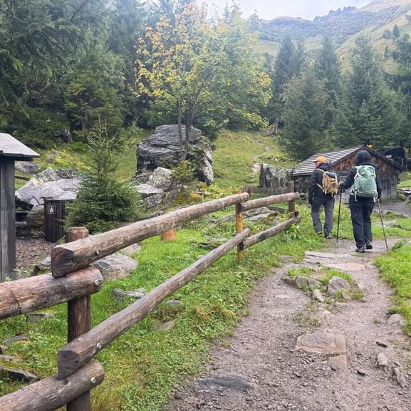 Zwei Wanderer mit Rucksäcken auf Bergweg vorbei an Holzzaun und Alphütten, umgeben von Wald und Bergen in der Ostschweiz.