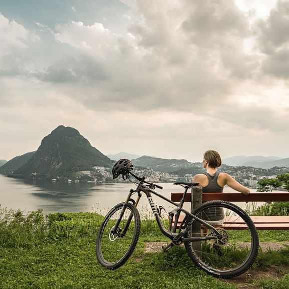 Eine Frau sitzt auf einer Bank mit Sicht auf den Lago di Lugano und den San Salvatore.