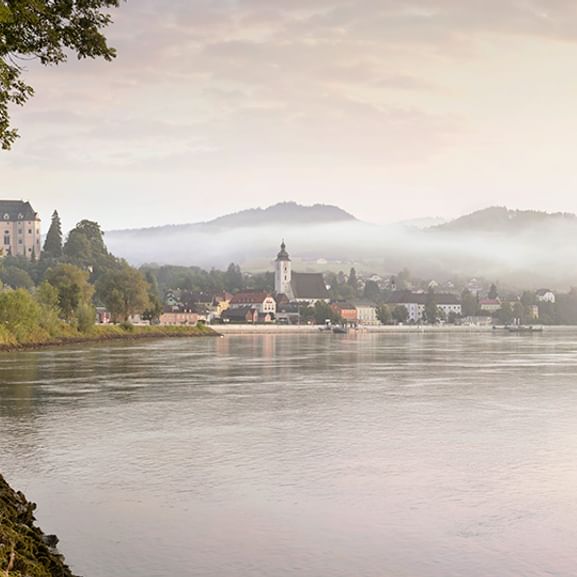 Drei Radfahrer fahren entlang des Donauradwegs bei Grein, Österreich zur goldenen Stunde mit nebligen Hügeln und Stadt im Hintergrund.