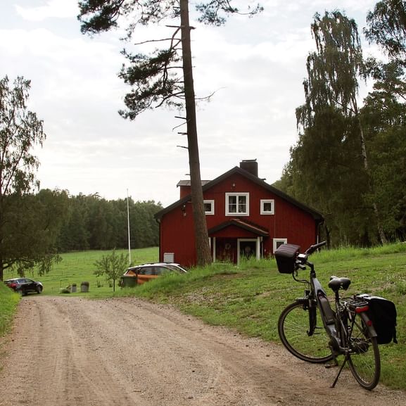 Tourenrad mit Taschen auf Schotterweg zu rotem Holzhaus umgeben von grünen Wiesen und Wald bei Stockholm.