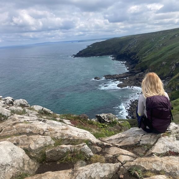 Wanderin mit lila Rucksack sitzt auf Felsenklippe in Cornwall und blickt auf türkisfarbenes Meer und grüne Küstenhügel unter bewölktem Himmel.