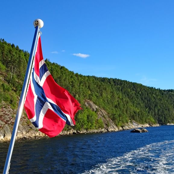 Drapeau norvégien flottant depuis un bateau sur l'Oslofjord avec collines boisées et côte rocheuse. Eau bleue et ciel clair en arrière-plan.