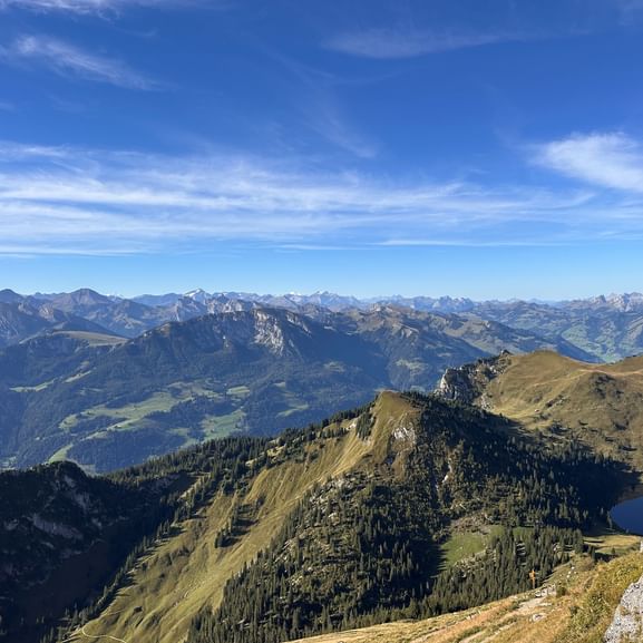 Panoramablick vom Stockhorn mit gestaffelten Bergketten, grünen Almwiesen, bewaldeten Hängen und einem kleinen Bergsee unter blauem Himmel.