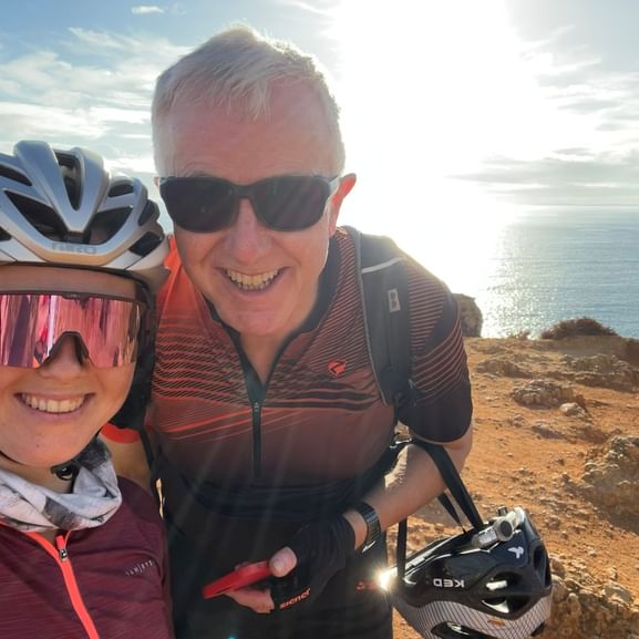 Zwei lächelnde Radfahrer in Radkleidung auf felsigen Küstenklippen nahe Ponte da Piedade, Algarve. Meer und blauer Himmel im Hintergrund.