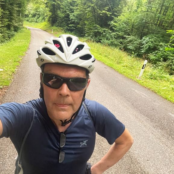 Radfahrer Werner macht Selfie auf Waldstraße mit weißem Helm und Sonnenbrille. Asphaltstraße führt durch üppigen grünen Wald.