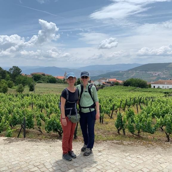 Zwei Wanderinnen mit Rucksäcken in Weinberg bei Vilarinho de São Romão. Grüne Reben, Hügel und weiße Gebäude unter blauem Himmel.