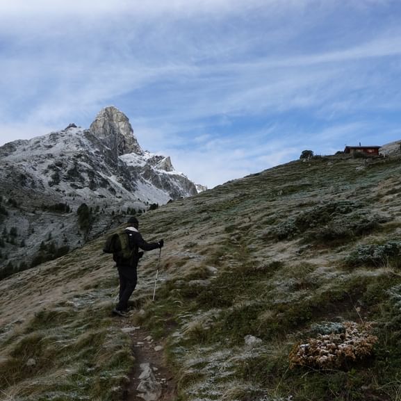 Wanderer mit Rucksack und Stöcken auf Alpenpfad mit verschneitem Meidhorn-Gipfel und Berghütte. Frost bedeckt das Gras entlang des Alpenpässewegs.
