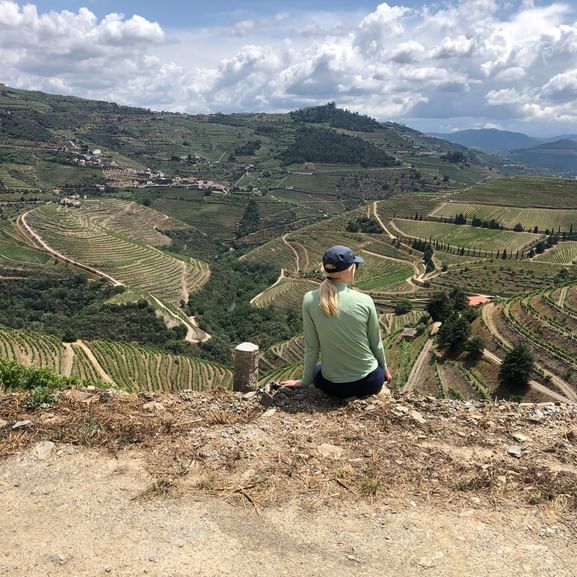 Wanderin sitzt auf Steinmauer mit Blick auf terrassierte Rebberge im Douro-Tal. Hügel mit Weinreihen unter bewölktem Himmel.