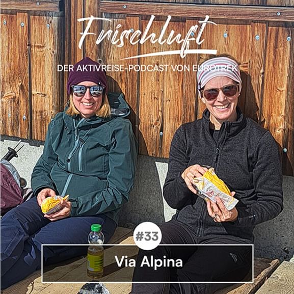 Zwei lächelnde Frauen in Outdoorkleidung sitzen mit Snacks auf einer Bank vor einer Holzhütte. Text: 'Forschaluft Folge 33 Via Alpina'.