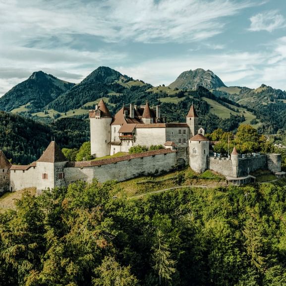 Schloss Greyerz thront auf einem bewaldeten Hügel mit dem Moléson-Gebirge im Hintergrund unter bewölktem Himmel.