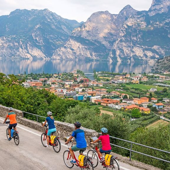 Groupe de cyclistes en maillots colorés arrêtés sur une route de montagne surplombant le lac de Garde avec des montagnes et une ville.